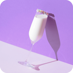 Champagne glass with sprinkles on the rim leans against a wall filled with vanilla protein shake
