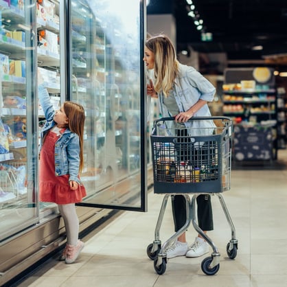 Woman pushes cart and holds open freezer door while young girl reaches up to shelf.